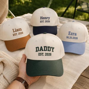 Puede incluir: Cuatro gorras de béisbol con coronas color blanco hueso y viseras de colores. Las gorras presentan texto bordado: Liam EST. 2026 con visera marrón, Henry EST. 2026 con visera azul marino, Ezra 06.20.2026 con visera azul claro y DADDY EST. 2026 con visera verde oscuro.