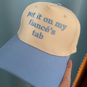 May include: A cream-coloured baseball cap with a light blue brim and matching light blue embroidered text that reads, "put it on my fiancé's tab." The cap features a structured front panel, a button on top, and stitched detailing on the brim.