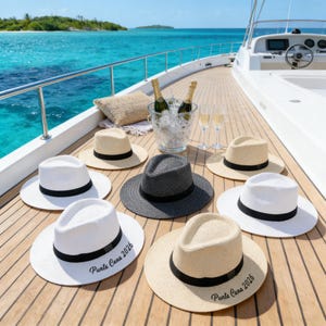 May include: Several fedora hats in white, beige, and grey with black bands are displayed on a yacht deck. Some hats are inscribed with "Punta Cana 2026". Champagne bottles are in an ice bucket, with two glasses beside them.