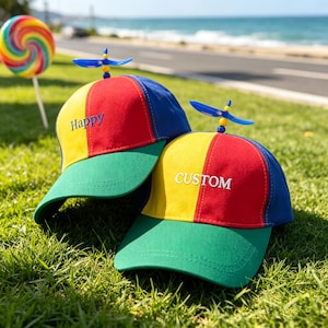 May include: Two multi-coloured baseball caps featuring red, yellow, blue, and green panels with a small blue propeller on top. One cap has the word Happy embroidered in blue, and the other has the word CUSTOM embroidered in white on the front.
