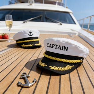 May include: Two white captain hats with black visors and gold trim rest on a wooden deck. One hat has an anchor emblem, the other reads "CAPTAIN". A silver anchor and a glass of champagne are also on the deck.