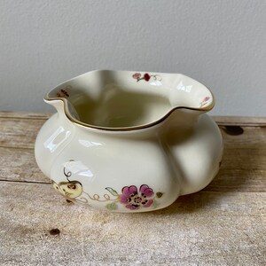 May include: A white ceramic bowl with a scalloped rim and floral design. The bowl has a gold trim and features pink, purple, and yellow flowers.