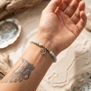 Peut inclure: Un bracelet de perles bleu clair avec des accents dorés et un charm en argent avec la lettre "m". Le bracelet est porté sur un poignet avec un tatouage d'ailes. Le fond comprend des coquillages et du bois flotté.