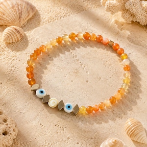 Peut inclure: Un bracelet de perles composé de pierres d'agate orange et jaune translucides, entrecoupées de petites entretoises métalliques en forme de losange, de deux perles œil bleu et d'une perle en coquillage blanc en spirale.