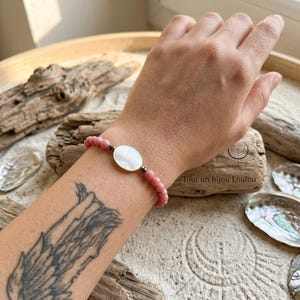 Peut inclure: Un bracelet de perles roses avec une pièce maîtresse ovale blanche est porté au poignet. Le bracelet est composé de petites perles roses rondes. La pièce maîtresse est de forme ovale blanche. Le poignet porte un tatouage d'aile d'ange. Le texte "Tout un bijou Loulou" est visible.