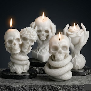 Puede incluir: Una colección de velas blancas con forma de calavera y mechas encendidas. Una vela presenta dos calaveras, otra tiene una calavera con las manos cubriendo su rostro, y otra tiene una calavera entrelazada con una serpiente. Las velas están sobre una superficie oscura.