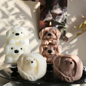 Puede incluir: Una colección de velas con forma de perro en varios colores. Una vela es una pila de dos perros blancos, otra es una pila de dos perros marrones con una mecha encendida, y también se ven otras dos velas de perro.