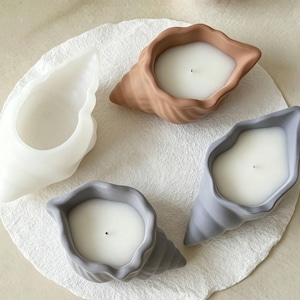 May include: Four seashell-shaped candles in various colors: white, tan, and two shades of gray. The candles are filled with white wax and have a single wick. The seashells are arranged on a white, textured surface.