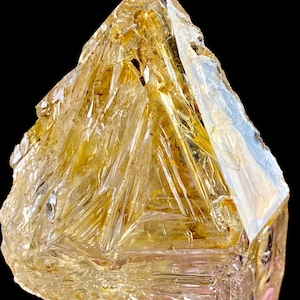 May include: A translucent, golden-yellow crystalline mineral specimen with a sharp, triangular-shaped termination. The surface features complex, intersecting geometric facets and internal striations that catch the light, creating a brilliant, multi-faceted appearance against a dark background.