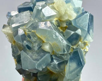 Może przedstawiać: Skupisko półprzezroczystych niebieskich kryształów o ostrych, geometrycznych fasetach i gładkich powierzchniach. Formacja mineralna charakteryzuje się różnymi odcieniami jasnego i głębokiego błękitu, przeplatanymi drobnymi plamami bladożółtej matrycy.
