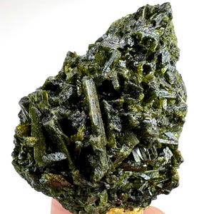 May include: A raw mineral specimen featuring a cluster of dark olive green, elongated, prismatic crystals with a vitreous lustre. The structure is dense and jagged, showcasing a complex arrangement of intersecting crystalline formations.