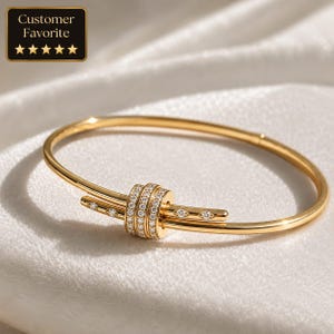Könnte beinhalten: Ein goldfarbenes Armband mit Scharnierdesign. Das Armband hat ein zentrales dekoratives Element mit drei Reihen kleiner, klarer Edelsteine. Zusätzliche Edelsteine sind entlang der Arme des Armbands gesetzt. Enthält ein "Customer Favorite"-Abzeichen.