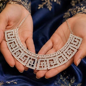 May include: A silver-toned statement necklace featuring a wide, curved collar design composed of repeating geometric Greek key patterns. The entire surface of the metal structure is encrusted with small, clear, sparkling stones.
