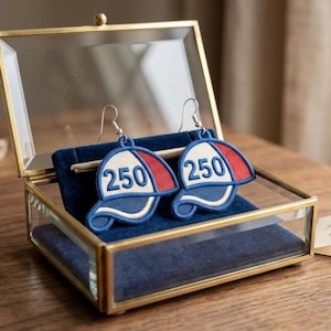 May include: A pair of baseball cap earrings with the number "250" embroidered on the front. The caps are white, red, and blue, with silver-colored ear wires. The earrings are displayed in a gold-framed glass box with a blue velvet interior.