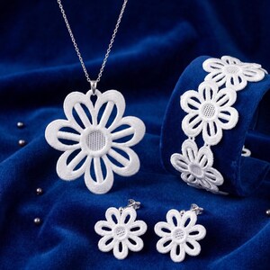Op de afbeelding: Een witte bloemen sieradenset op een blauw fluwelen oppervlak. De set bevat een ketting met een grote bloemhanger, een armband en een paar oorbellen. De bloemmotieven hebben een gedetailleerd, opengewerkt patroon.