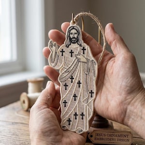 May include: Embroidered ornament featuring a depiction of Jesus with a halo and long hair, adorned with black crosses. The ornament is cream-colored with a brown twine loop for hanging. The text "JESUS ORNAMENT EMBROIDERY DESIGN" is on a small wooden tag.