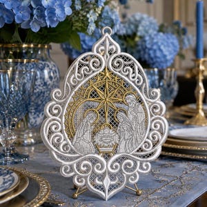 May include: A white and gold embroidered freestanding lace ornament featuring a nativity scene with a central star, Mary, Joseph, and the infant Jesus in a manger, all enclosed within an intricate, swirling, filigree-style decorative frame.