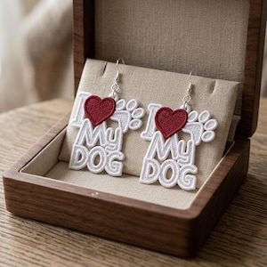 May include: White embroidered earrings with the text "I Love My Dog" and a red heart. The earrings are displayed in a wooden jewelry box with a beige interior. The earrings are a dog-themed accessory.
