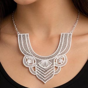 May include: A silver-toned statement necklace featuring an intricate filigree design with a wide, U-shaped collar. The piece displays delicate, lace-like metalwork patterns, including fan-shaped motifs and geometric lines, suspended from a simple chain.
