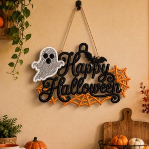 May include: A hanging wall sign featuring the black embroidered text Happy Halloween, a white ghost with a black text pattern, a small black bat, and orange spider webs. The sign is suspended by a tan twine cord from a black wall hook.