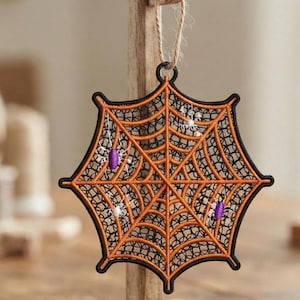 May include: A decorative hanging ornament featuring a black and orange embroidered spiderweb design. The web is accented with small purple and white thread spiders and is suspended by a thin tan twine loop attached to the top of the structure.