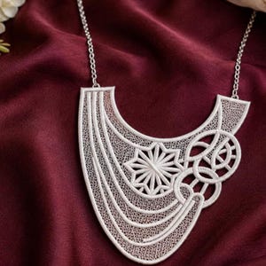 May include: A white lace statement necklace featuring an intricate, asymmetrical design with a central starburst motif, circular patterns, and flowing curved lines. The piece is suspended from a delicate silver-toned metal chain.