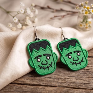 May include: A pair of green Frankenstein monster earrings with black hair and stitched details. The earrings are embroidered with a smiling face and a stitched mouth. They hang from silver-colored hooks.