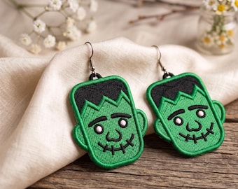 Puede incluir: Un par de pendientes verdes de Frankenstein con pelo negro y detalles cosidos. Los pendientes están bordados con una cara sonriente y una boca cosida. Cuelgan de ganchos de color plateado.