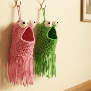 May include: Two handmade crochet wall organizers, one pink and one green, each resembling a whimsical monster with large white and black eyes. The organizers have open mouths and fringed bottoms, hanging from hooks on a beige wall.