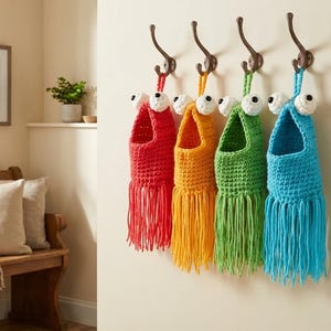 May include: Five colorful, handmade crochet wall pockets hang from bronze hooks. Each pocket is a different color: red, orange, green, and blue. They have white and black googly eyes and fringe.