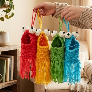 May include: Four colorful, handmade crochet monster pouches with dangling fringe and large googly eyes are held up. The pouches are red, yellow, green, and blue. The background includes a wooden shelf with plants and books.