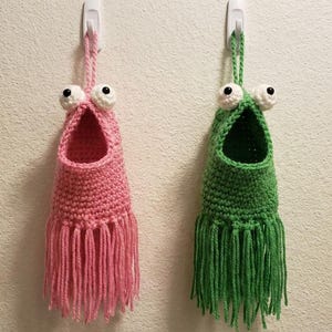 May include: Two handmade crochet wall organizers, one pink and one green, each with large googly eyes and a fringed bottom. They are hanging on a white wall, adding a whimsical touch to any room.