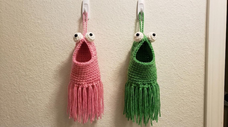 May include: Two handmade crochet wall organizers, one pink and one green, each with large googly eyes and a fringed bottom. They are hanging on a white wall, adding a whimsical touch to any room.