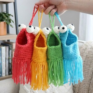 May include: Four colorful, handmade crochet monster pouches are held up. Each pouch is a different color: red, yellow, green, and blue. They have white and black googly eyes and fringe at the bottom. The pouches are hanging from a loop.