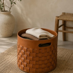 May include: A round, woven leather storage basket in a warm brown hue. The basket contains folded beige and brown textiles. The basket sits on a textured, neutral-toned rug. A decorative vase with greenery is in the background.