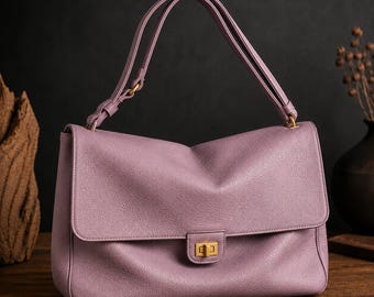 Könnte beinhalten: Eine lavendelfarbene Handtasche mit strukturierter Oberfläche, die über einen rechteckigen Klappenverschluss mit einem goldfarbenen Drehverschluss aus Metall verfügt. Die Tasche hat einen passenden Schulterriemen mit goldfarbenen Beschlägen.