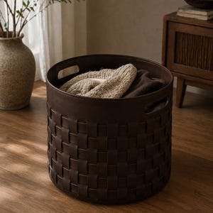 May include: A dark brown woven leather storage basket with two handles. The basket is filled with folded beige and brown textiles. The basket is sitting on a wooden floor. Home decor.