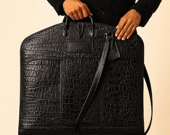 Könnte beinhalten: Schwarze Kleiderschutzhülle aus Leder mit Krokodiltextur. Die Tasche hat einen oberen Griff, einen Schultergurt und ein rechteckiges Etikett. Die Tasche wird von einer Person gehalten, die eine schwarze Jacke trägt.