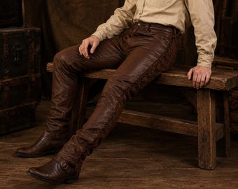 May include: Dark brown leather pants featuring a decorative lace-up detail running down the outer side of each leg. The pants have a fitted silhouette and are paired with dark brown leather boots that include a buckle strap across the ankle.