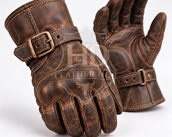Könnte beinhalten: Ein Paar Handschuhe aus braunem, abgenutztem Leder mit einem Handgelenkriemen und einer messingfarbenen Metallschnalle. Die Handschuhe haben verstärkte Knöchelpolster und sichtbare Nähte. Der Text LEATHER HUB ist zu sehen.
