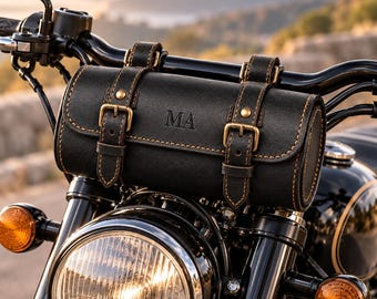 Könnte beinhalten: Eine schwarze, zylindrische Motorrad-Lenkertasche mit zwei vertikalen Riemen, messingfarbenen Schnallen und sichtbaren Nähten. In der Mitte der Tasche sind die Buchstaben MA eingeprägt. Die Tasche ist am schwarzen Lenker eines Motorrads befestigt.