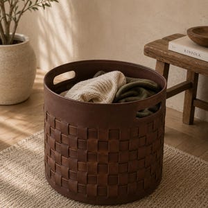 May include: A brown woven leather laundry basket with two handles, filled with folded sweaters. The basket sits on a beige rug, with a plant and wooden stool in the background. The basket's design features a repeating square pattern.