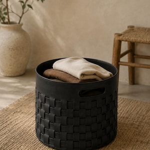 May include: A black woven storage basket with a round shape, holding folded beige and brown textiles. The basket sits on a natural fiber rug, with a neutral background and a decorative vase.