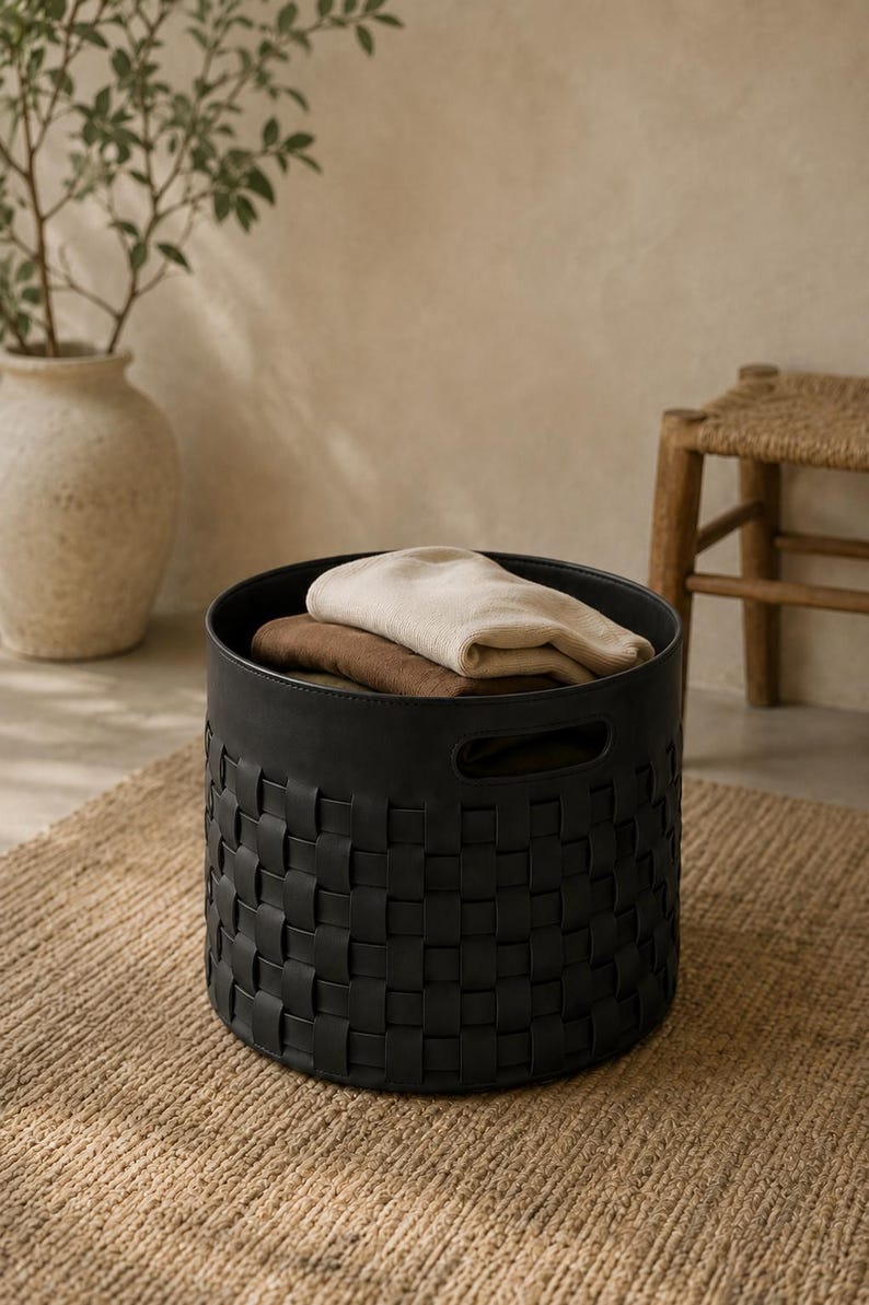 May include: A black woven storage basket with a round shape, holding folded beige and brown textiles. The basket sits on a natural fiber rug, with a neutral background and a decorative vase.