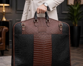 Könnte beinhalten: Eine große Kleidersacktasche mit einer strukturierten Krokodilprägung in abwechselnden schwarzen und braunen Feldern. Die Tasche verfügt über zwei schwarze Tragegriffe, goldfarbene Metallschnallen an den oberen Ecken und ein braunes Lederetikett.