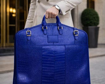 Könnte beinhalten: Eine leuchtend königsblaue Kleidersacktasche mit einer strukturierten Krokodilleder-Optik. Die Tasche verfügt über zwei Tragegriffe, goldfarbene Metallschnallen an den Seitengurten und ein rechteckiges, leeres Etikett in der Mitte der Vorderseite.