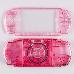 May include: A disassembled, transparent pink Sony PSP handheld gaming console. The top and bottom halves are separated, revealing internal components. The Sony logo is visible on the top half. The bottom half has the UMD logo.
