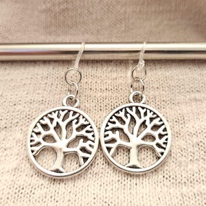 May include: A pair of silver-colored earrings featuring a tree of life design. Each earring has a circular pendant with a detailed tree silhouette. The earrings hang from a silver-colored hook. The background is a light beige fabric.