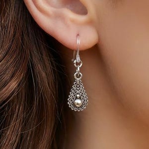 May include: A silver teardrop earring with a detailed filigree design and a small ball at the bottom. The earring is suspended from a simple hook. The jewelry is shown against a blurred background.