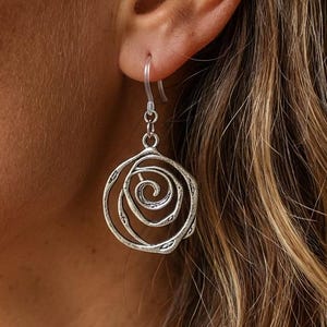 May include: Silver-toned spiral earrings. The earrings feature a circular design with a spiral center and a hook closure. The earrings are shown against a blurred background of skin and hair.